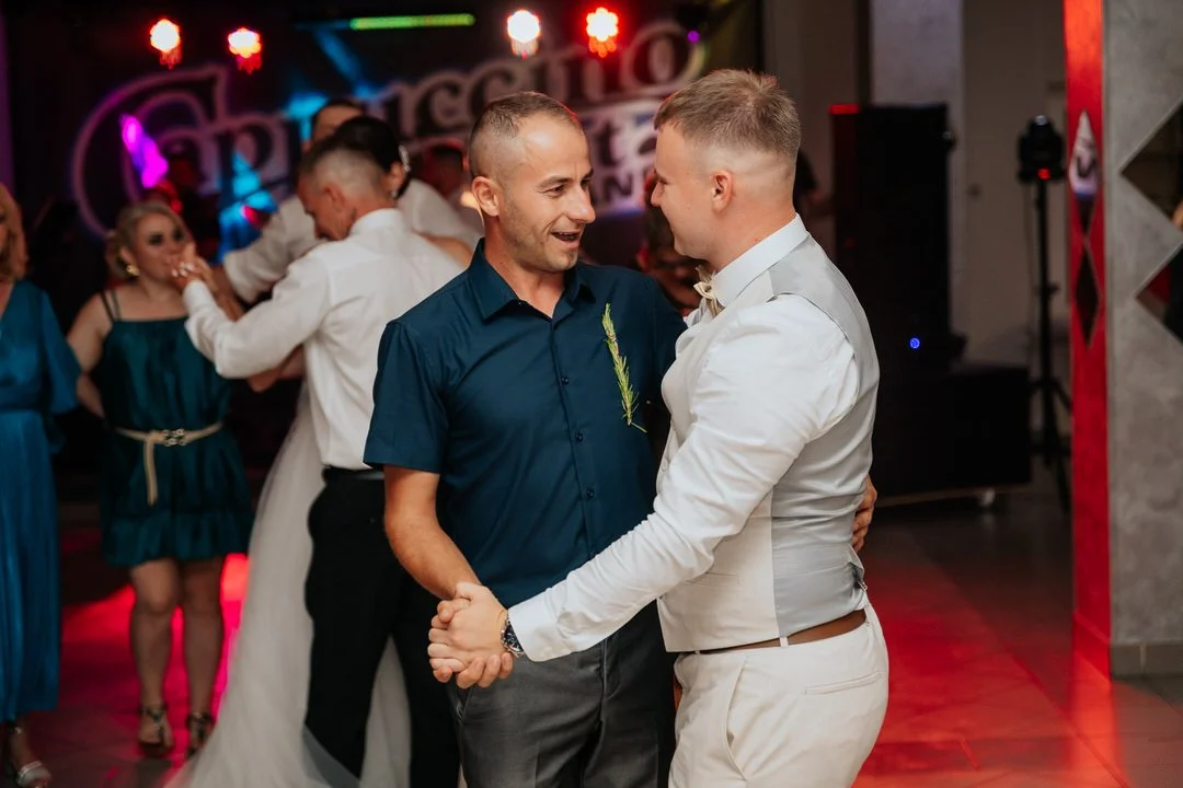 Two men dance and hold hands at a celebration, with other people dancing and socializing in the background, colorful lights, and a banner that reads 'Perfecto'.