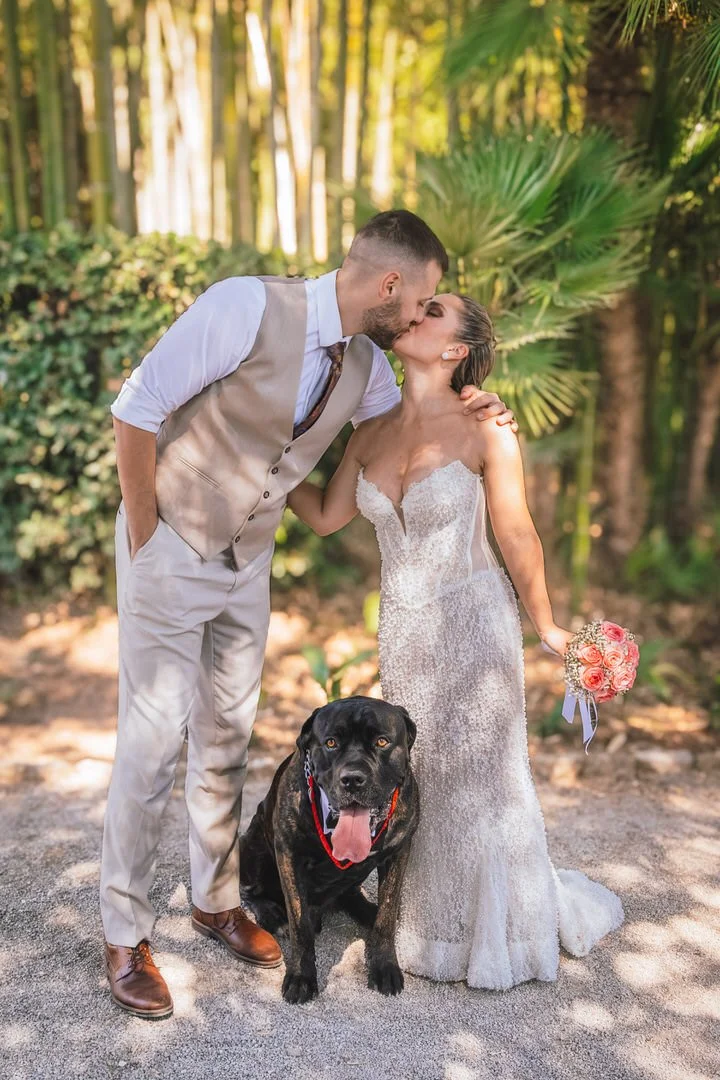 Newlywed couple sharing a kiss with their large dog in Park Angiolina, Opatija, Kvarner, surrounded by woodland and sunlight filtering through the trees.