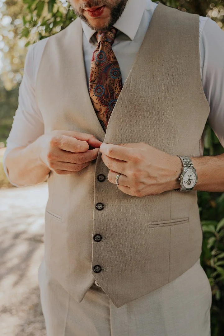 A man wearing a light-colored vest and matching pants buttoning his vest, with a white dress shirt and a paisley tie, outdoors during daytime.