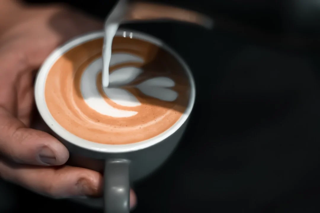 A person creates latte art in a cup of coffee with steamed milk. The design includes hearts and swirls.