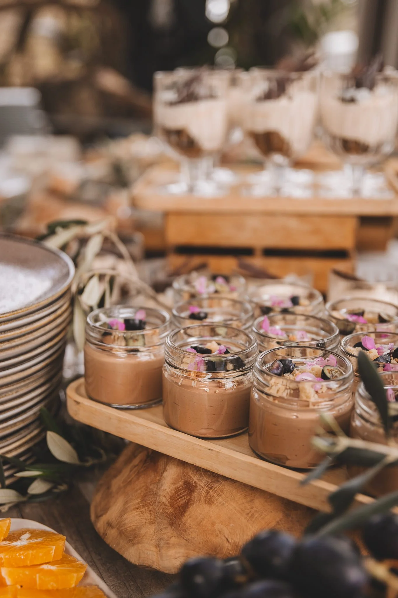 Various desserts and toppings displayed on a wooden table at a buffet or outdoor event, including small jars of mousse with pink flower petals and a chocolate bar from a San Canzian Istria brunch in Croatia.