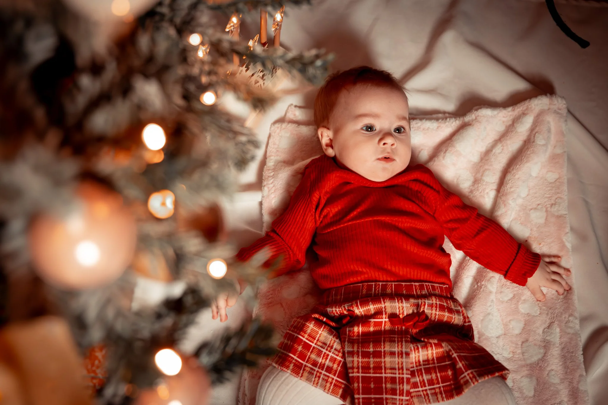 A baby lying on a blanket under a Christmas tree decorated with lights, wearing red holiday clothing.
