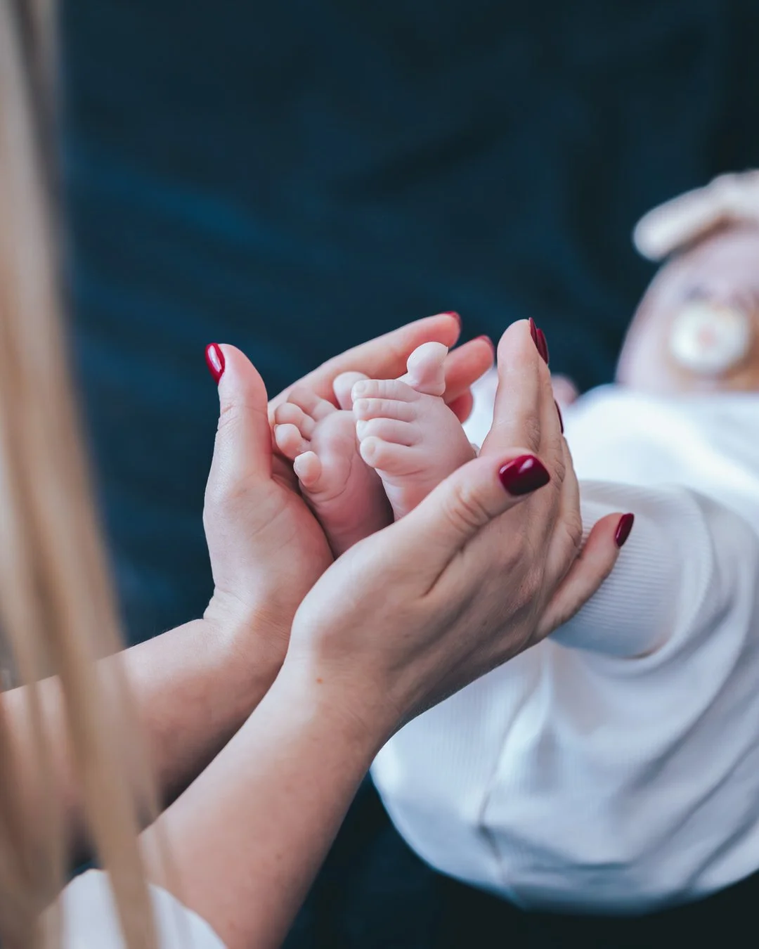 An adult woman gently holding a newborn baby's tiny hand in her palm by a window with soft natural light.