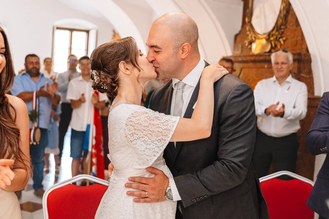 Bride and groom sharing a romantic kiss during their wedding ceremony at Eltz Castle in Vukovar, Croatia, surrounded by cheering friends and family.