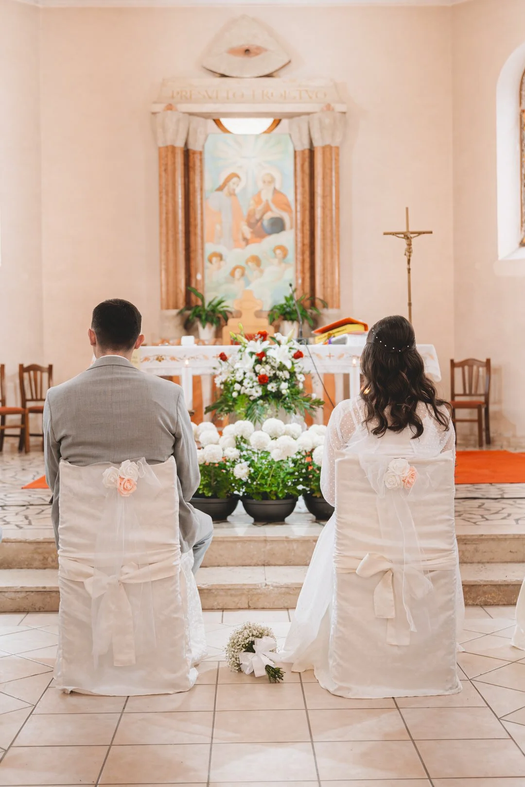 A bride and groom sitting in a church during their wedding ceremony, facing the altar decorated with flowers and religious artwork.