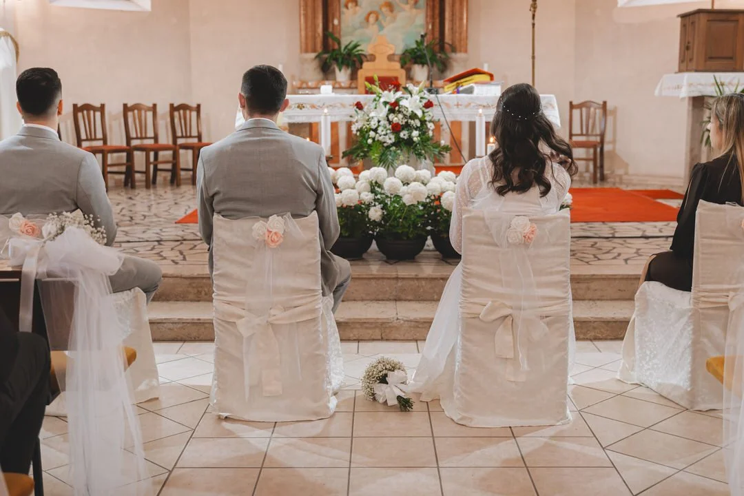 A wedding ceremony in a church with four people seated facing the altar, decorated with white flowers and chairs covered in white fabric with pink roses.