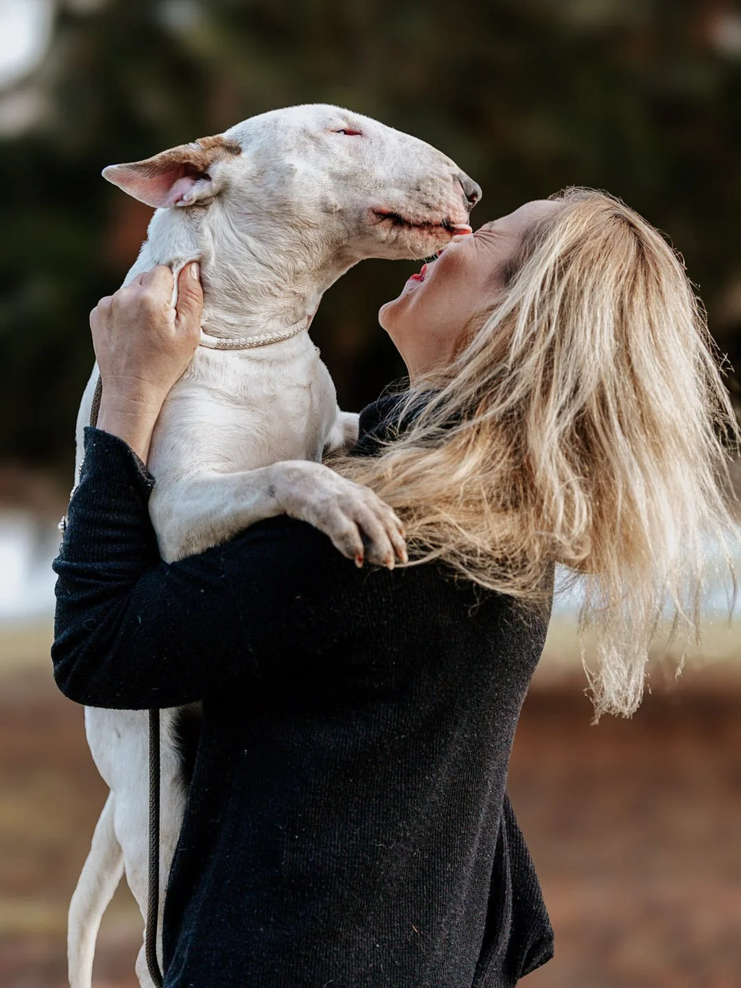 A woman holding a white dog while kissing it on the face outdoors.