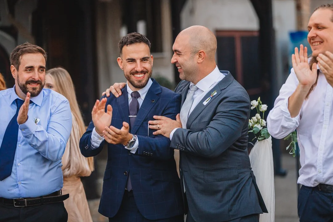 Group of men in suits at a celebration, smiling and clapping. One man is helping another adjust his jacket, indicating joy and camaraderie.