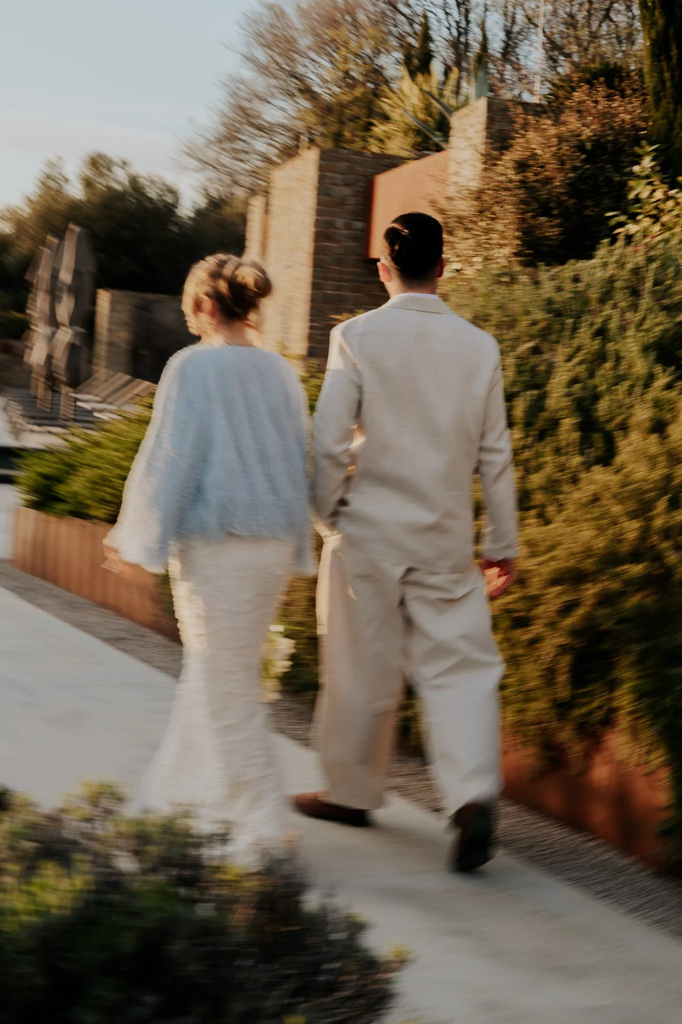 A couple dressed in white walking away from the camera on a paved path in a garden with trees and shrubs.