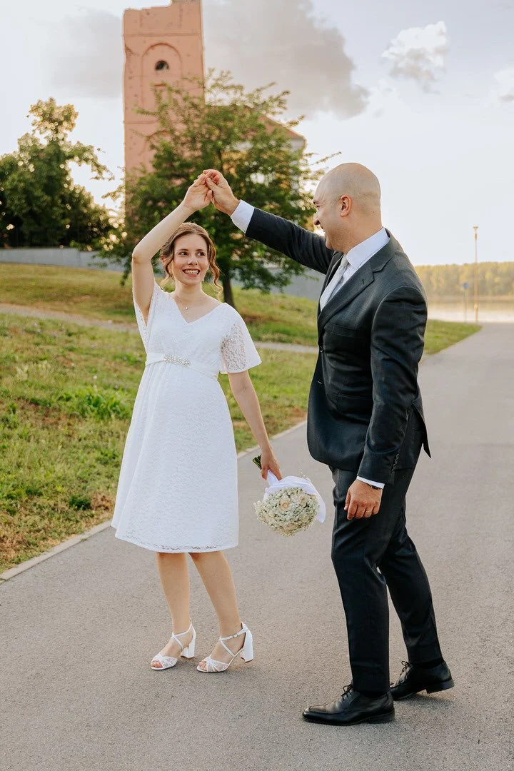 Bride and groom dancing outdoors during sunset, with a church in the background. The bride is wearing a white dress and holding a bouquet, while the groom is in a black suit.