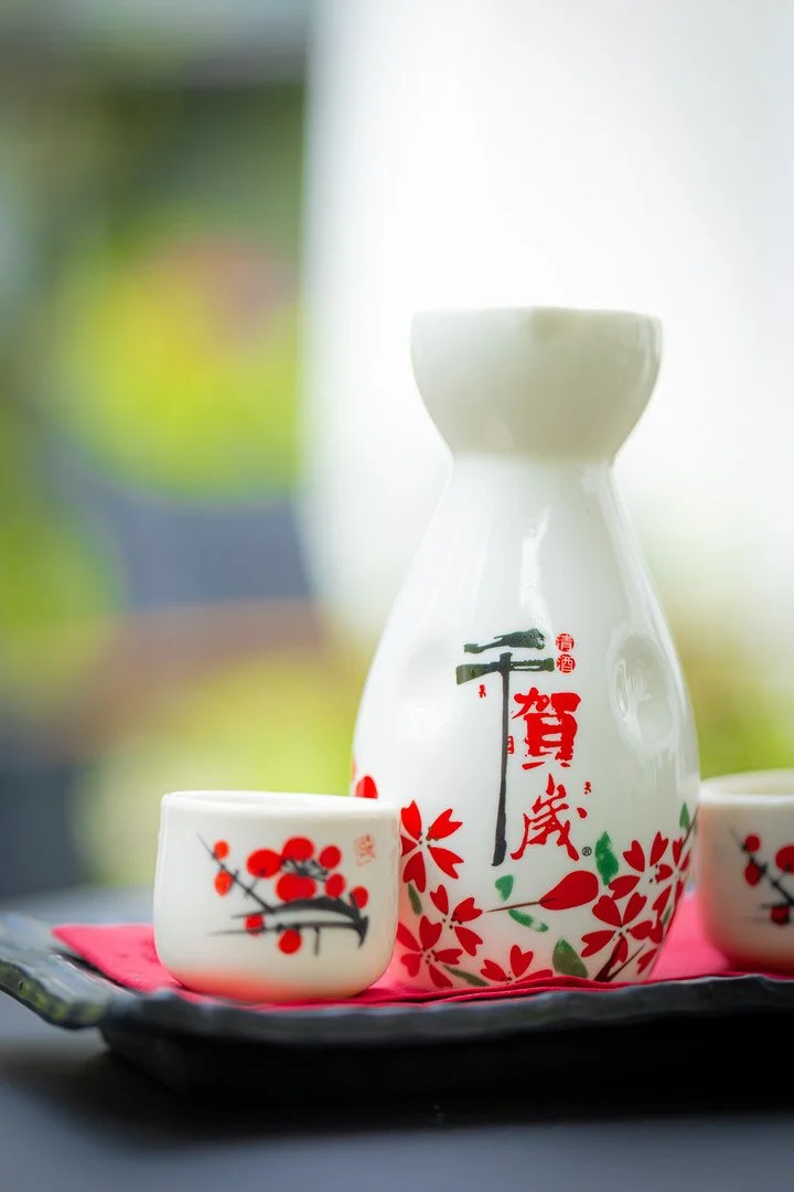 A white ceramic sake set with red and green floral and Japanese calligraphy designs, placed on a black tray with a pink cloth, with a blurred outdoor background.