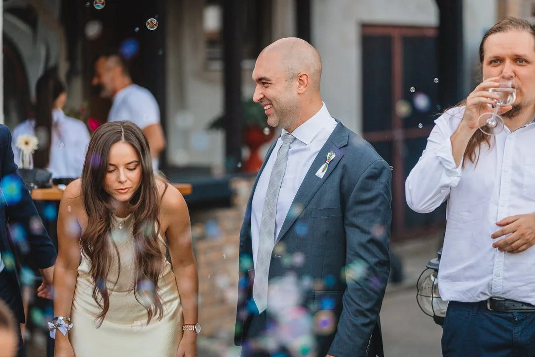 People at a social event, with a woman in a cream dress looking down, a man in a suit smiling, and another man drinking from a glass, with blurred background of a restaurant or bar