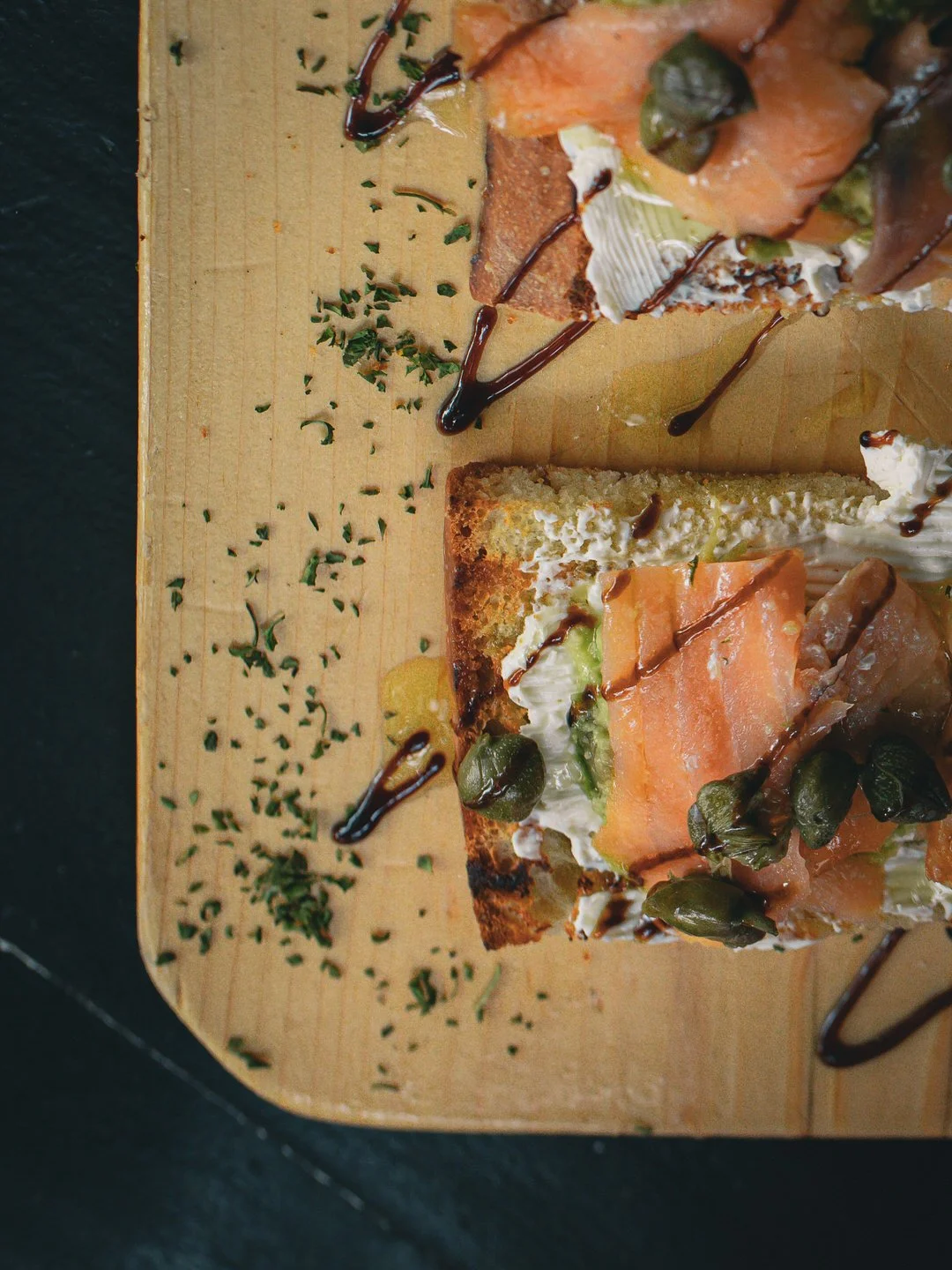 Slices of smoked salmon and grilled bread topped with capers, served on wooden cutting board with drizzled balsamic glaze and sprinkled herbs.