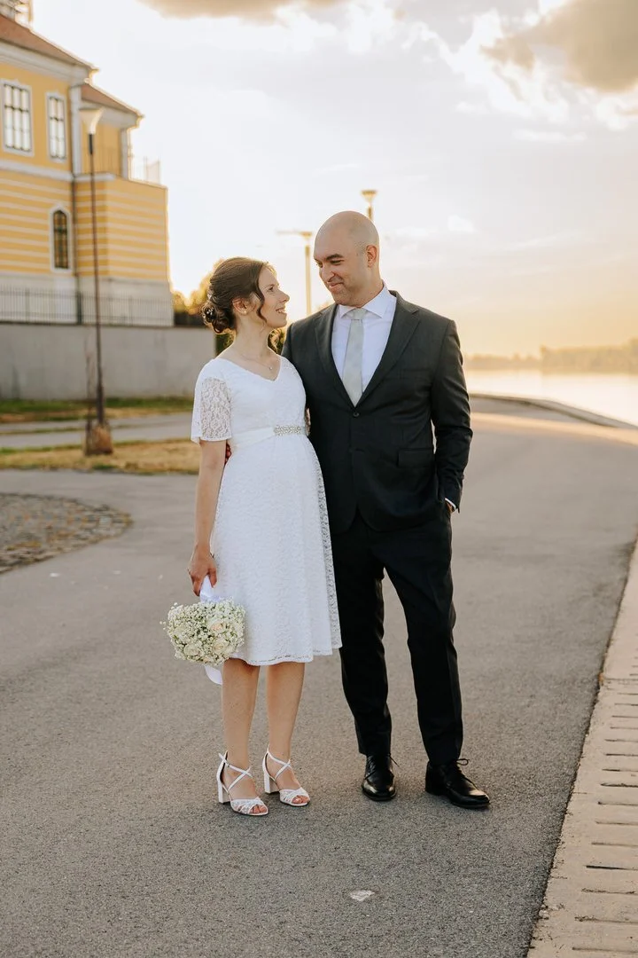 A bride and groom standing together outdoors near a waterfront during sunset. The bride is wearing a white lace dress and holding a small bouquet of white flowers, while the groom is dressed in a black suit with a white shirt and tie.