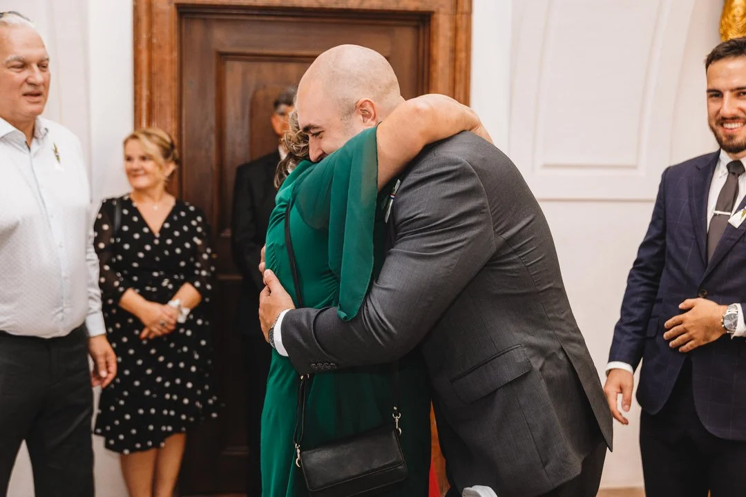 A man and woman hugging in a formal indoor setting, surrounded by several smiling people, including a woman in a polka dot dress and two men in suits.