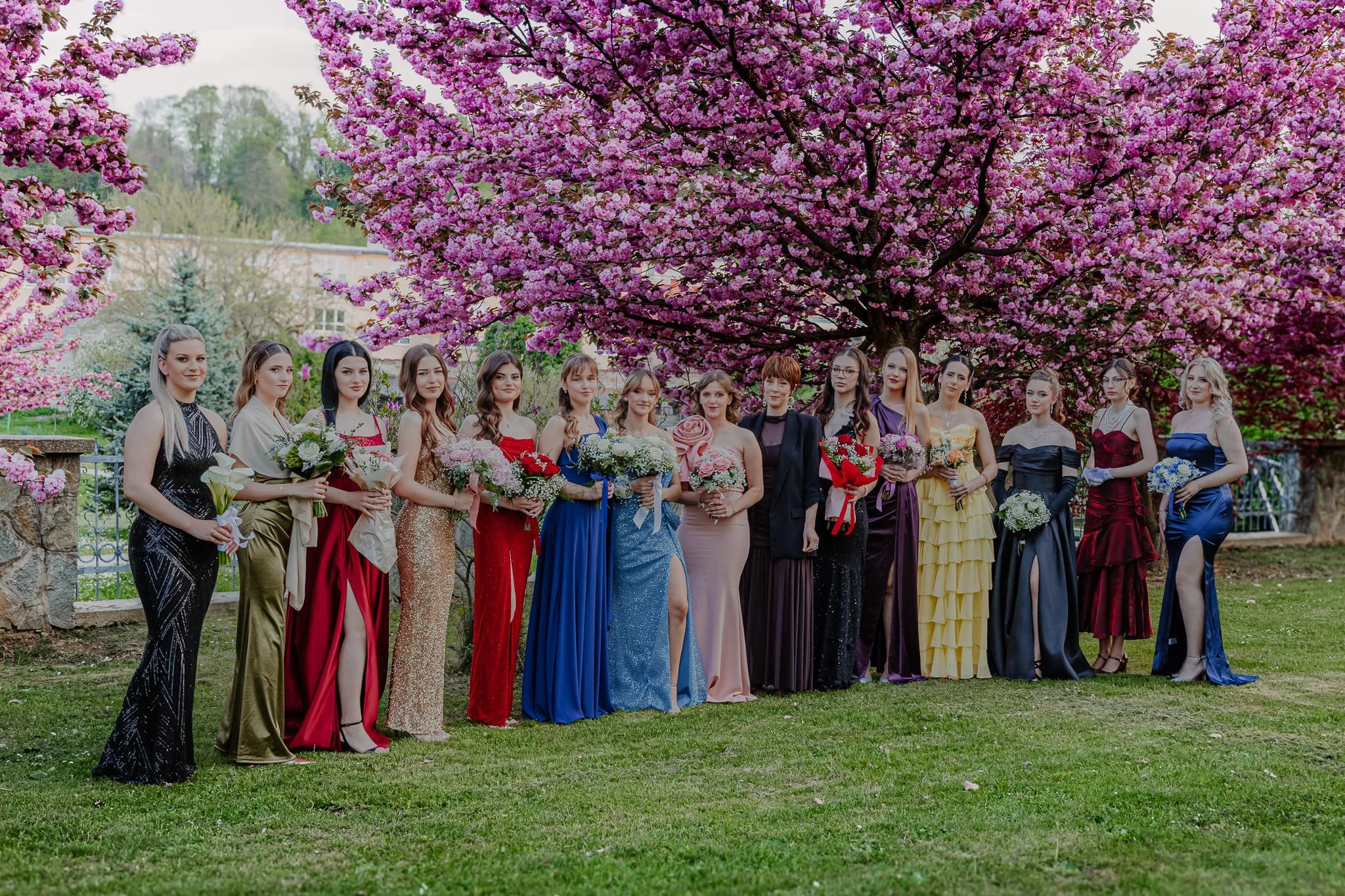 A group of women in colorful dresses standing outdoors under a pink flowering tree, holding bouquets of flowers at their prom