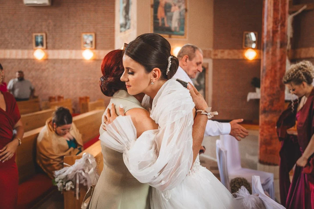 Two women hugging inside a church during a wedding ceremony, with guests seated and standing around.