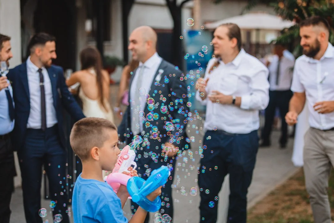 A young boy is blowing bubbles with a bubble gun at an outdoor gathering, with adults in semi-formal attire chatting and smiling in the background.
