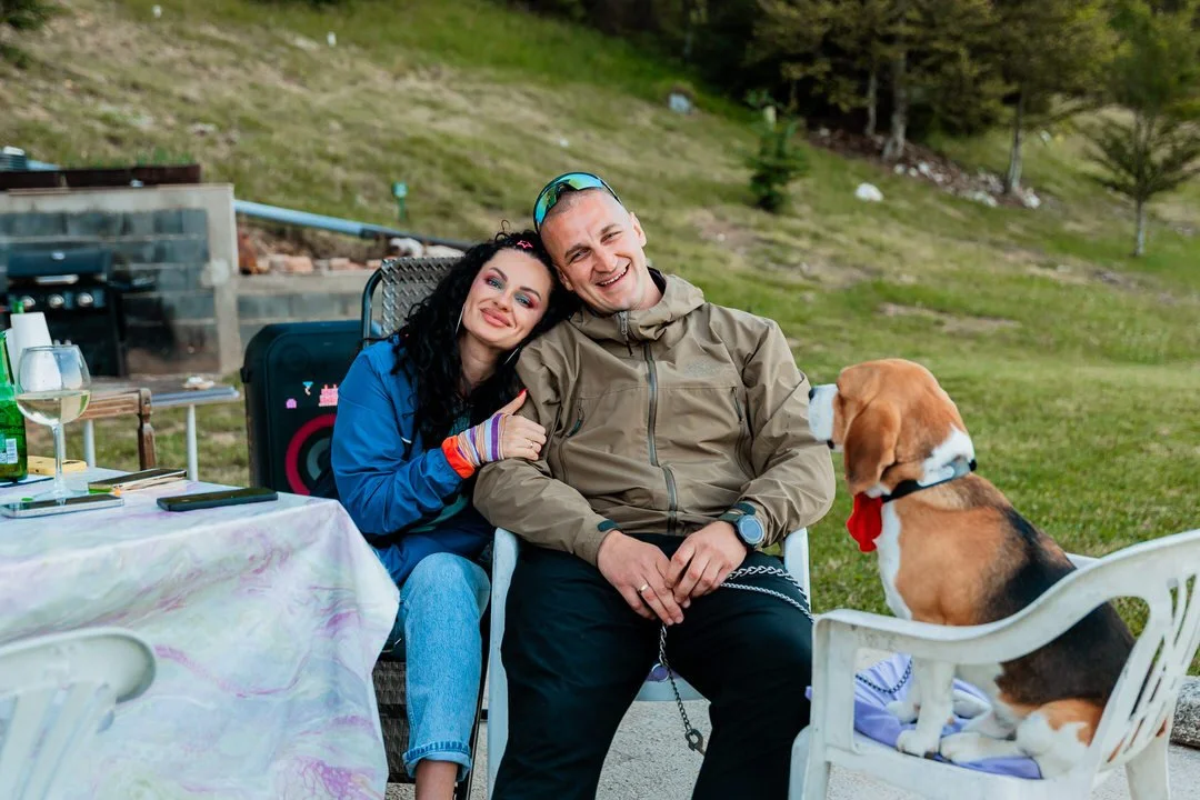 A couple sitting outdoors, smiling, with a dog wearing a red bow tie, on a patio with a grassy hillside in the background.