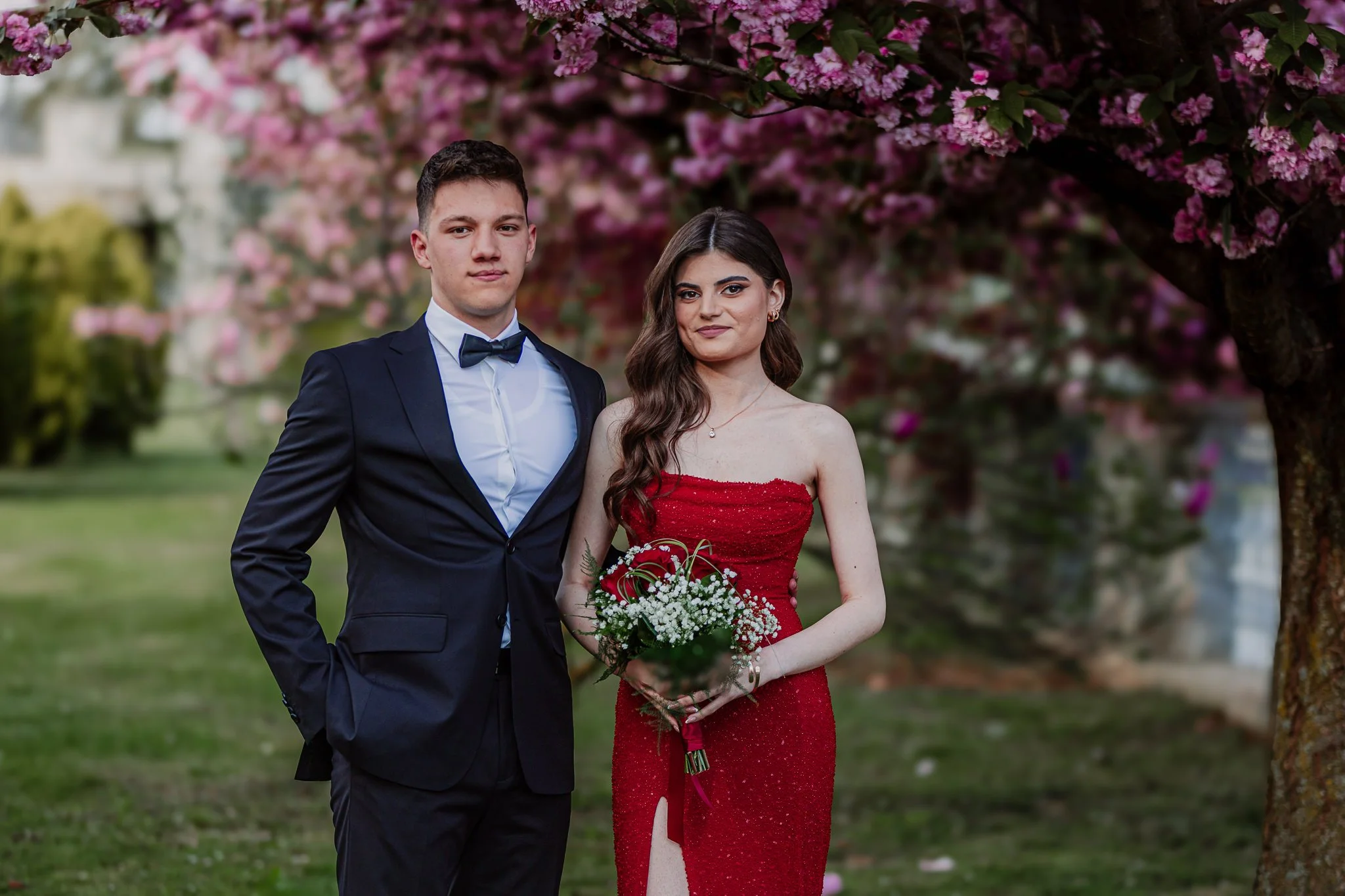 A young man in a navy tuxedo and a young woman in a red dress holding a bouquet of white flowers standing outdoors in front of pink flowering trees.