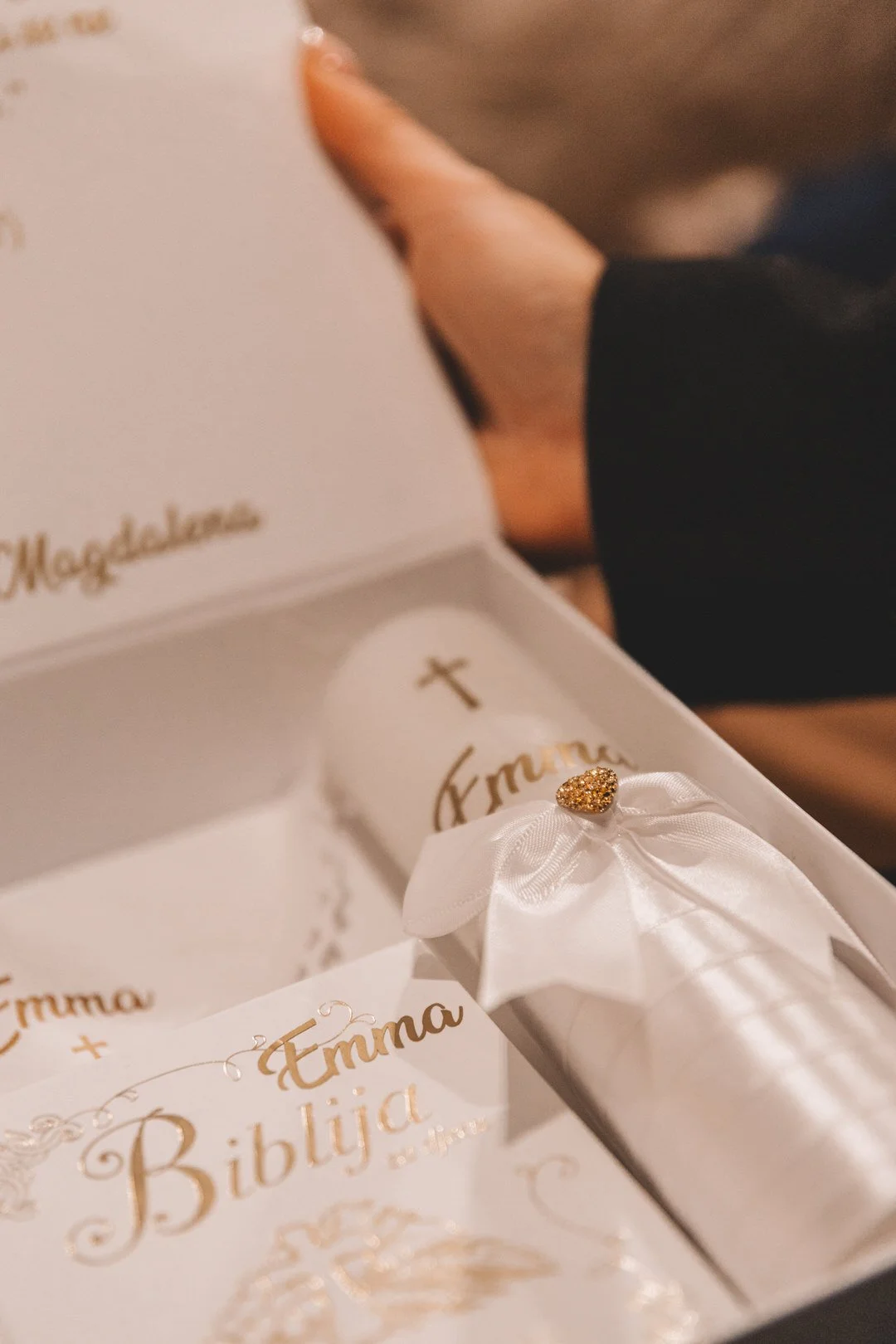 A close-up of a white box with gold text and decorations, containing a white ribbon with a small gold heart, a cross, and a card that reads 'Emma' in gold lettering.