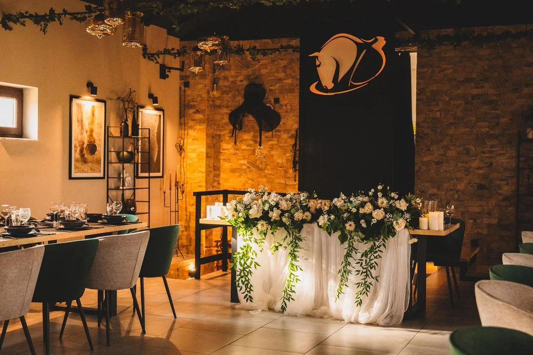 Elegant indoor wedding reception space at Dunavski Raj estate, featuring a decorated table with a floral centerpiece, candles, a black curtain with a horse head and number two logo, brick walls, artwork, and chairs around the tables.
