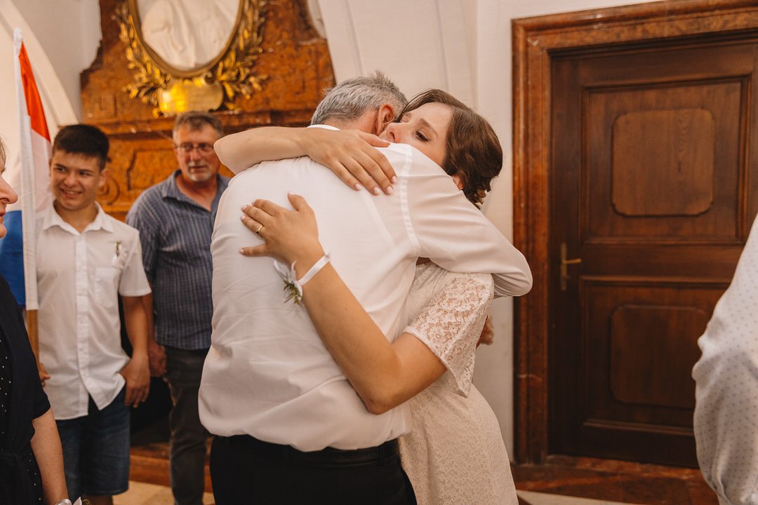 A woman hugging an older man at a formal gathering, with other people smiling and watching in a warmly decorated room.