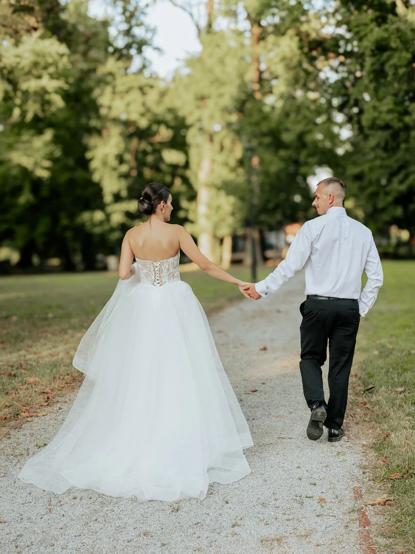 A bride in a white wedding gown and a groom in a white shirt and black pants walk hand in hand outdoors on a gravel path in a park with green trees.