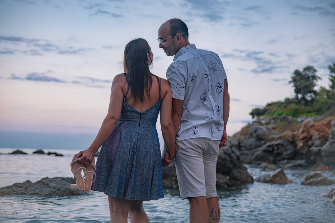 A couple holding hands on a rocky beach at sunset on Krk island croatia, the woman holding sandals, both looking at each other.