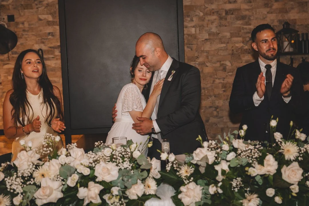 A wedding reception with a bride and groom embracing at the head table, surrounded by floral arrangements. Two other guests are clapping and watching the couple.