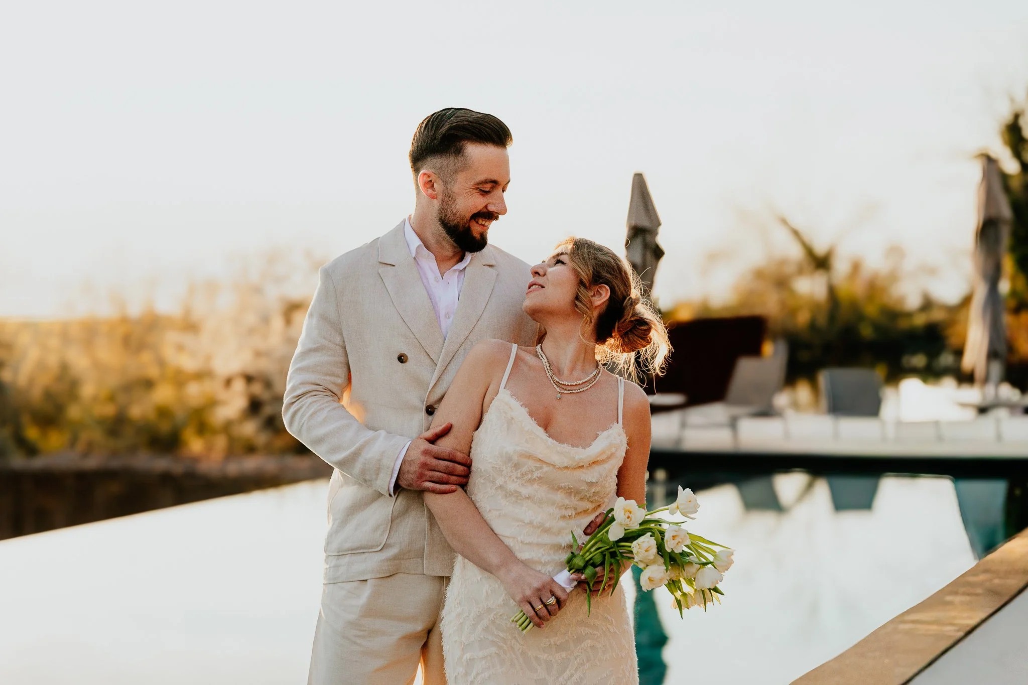 A wedding couple stands outdoors near a body of water, sharing a joyful moment during sunset. The groom, dressed in a light-colored suit, looks lovingly at the bride. The bride, in a lace wedding dress, holds a bouquet of white flowers.