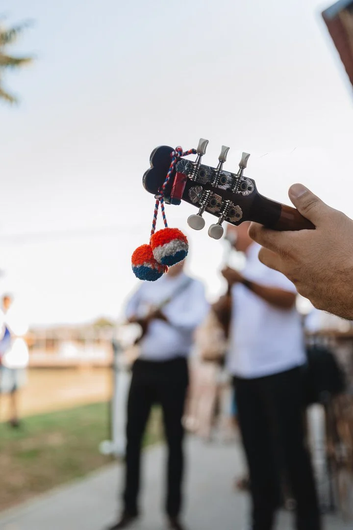 A person's hand holding a tambura headstock with decorative pom-poms hanging from it, with a blurred group of people in the background outdoors.