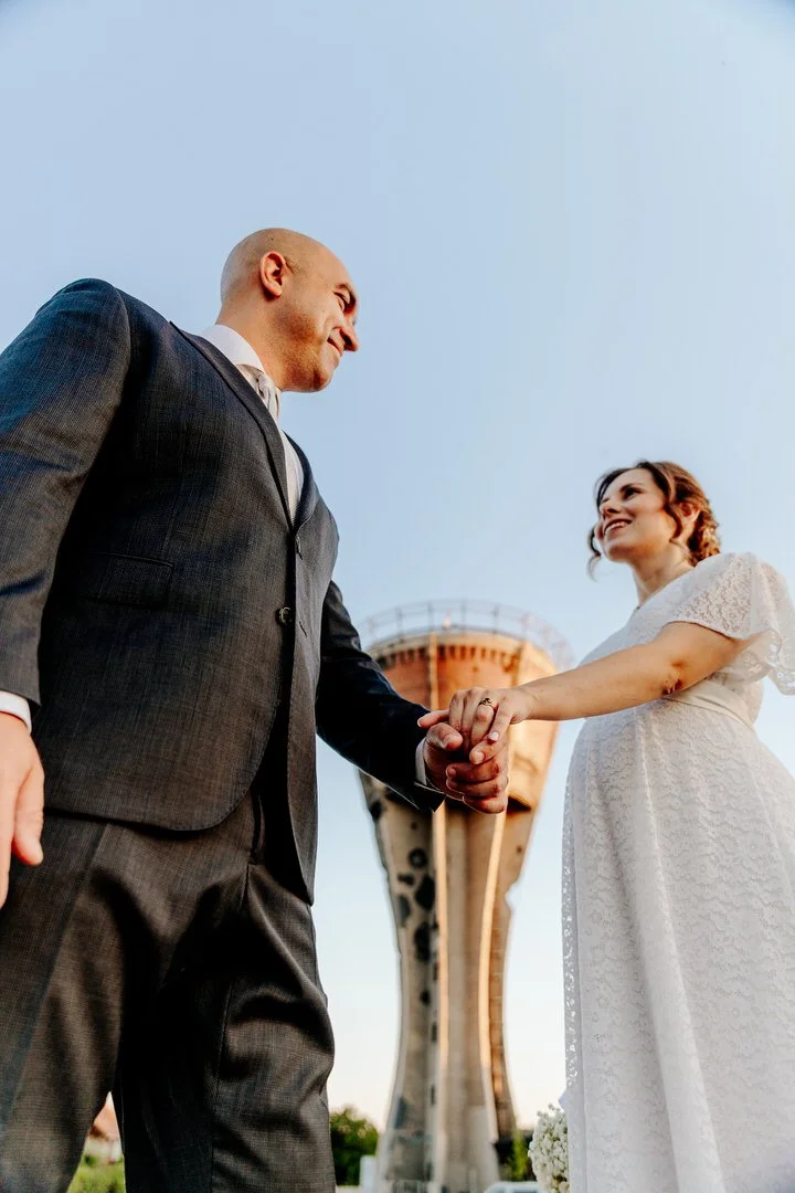 Bride and groom holding hands and smiling at each other outdoors, with the Vukovar Water Tower in the background during daytime.