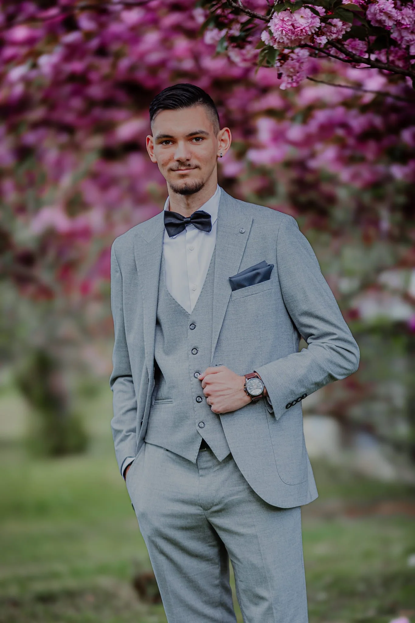 a high school graduate wearing a light gray suit, white shirt, and black bow tie stands outdoors in front of blooming pink cherry blossom trees.