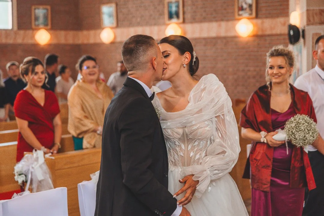 A bride and groom share a kiss during their wedding ceremony in a church, with guests watching and smiling in the background.