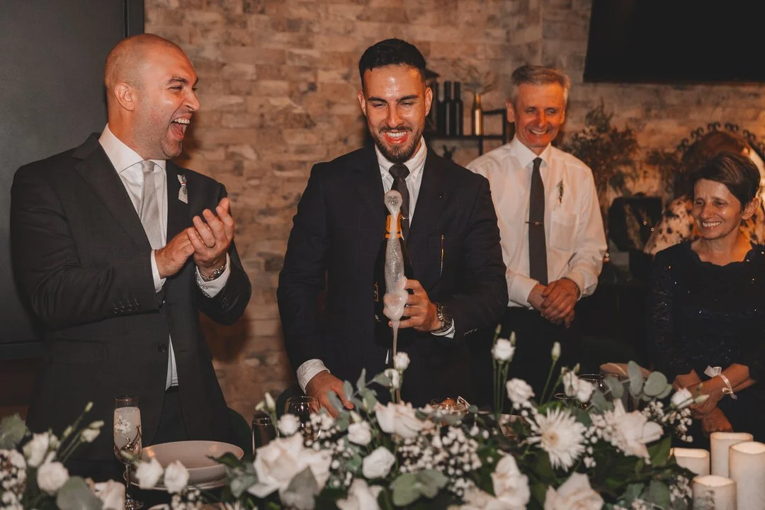 Group of five people at a celebration, with one person opening a bottle of champagne and foam spilling out, surrounded by floral arrangements and candles.