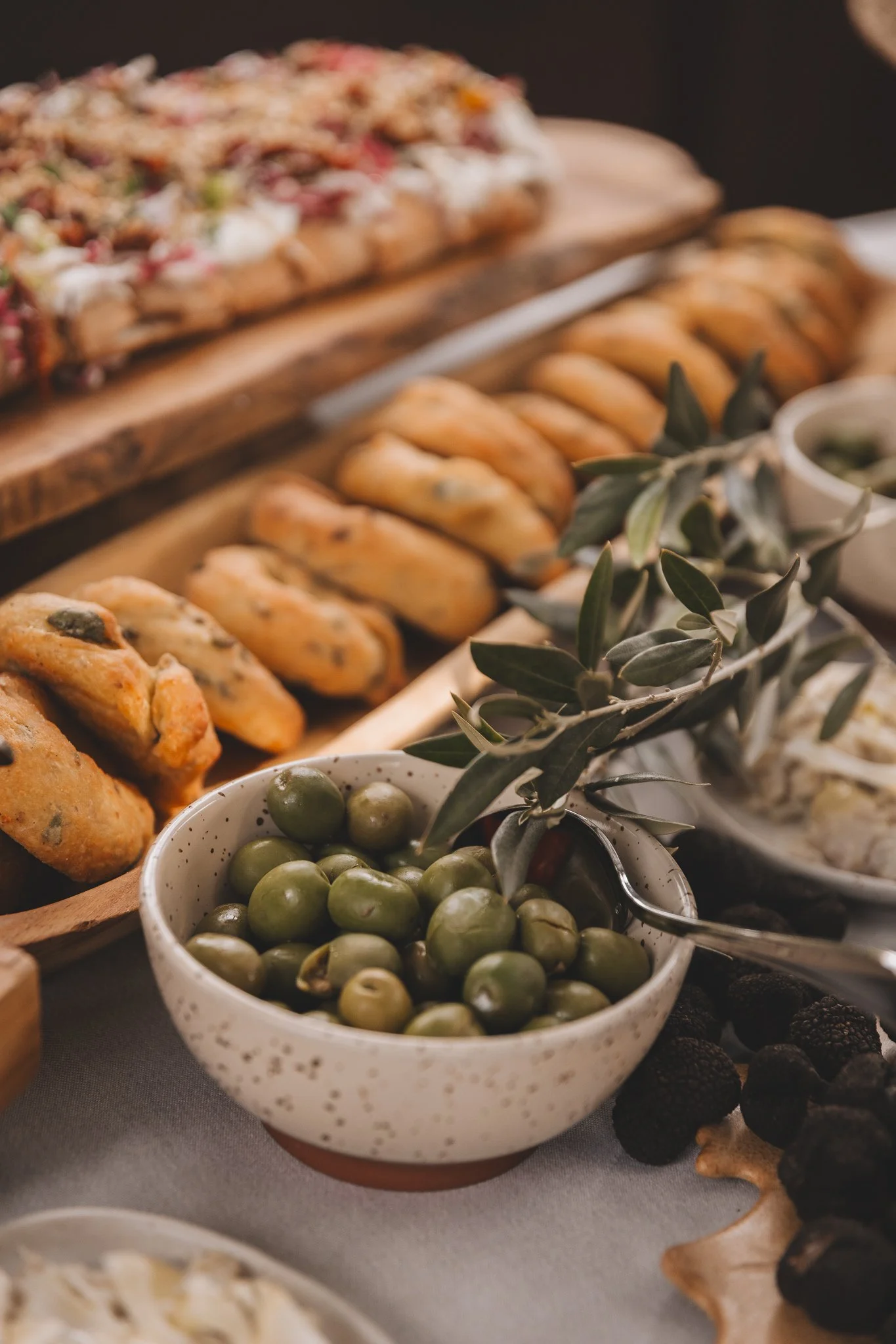 Bowl of green olives with olive leaves in the foreground, assorted bread, and a charcuterie board with various cheeses and meats in the background from a San Canzian Istria brunch in Croatia.