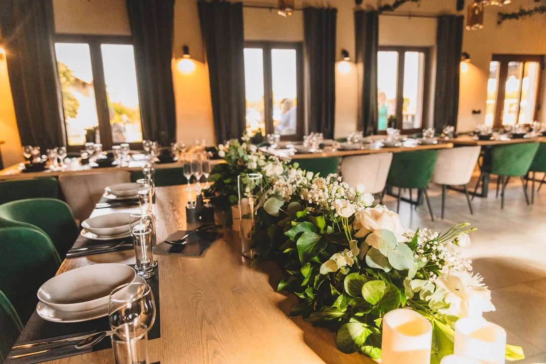 Elegant event space with decorated tables, floral centerpiece, candles, and large windows with dark curtains.