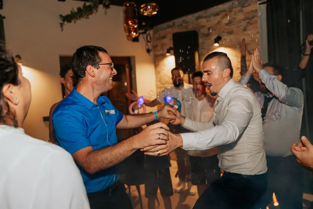 Group of people dancing and celebrating indoors at a party or wedding, with some holding hands and others in the background clapping and enjoying the moment.