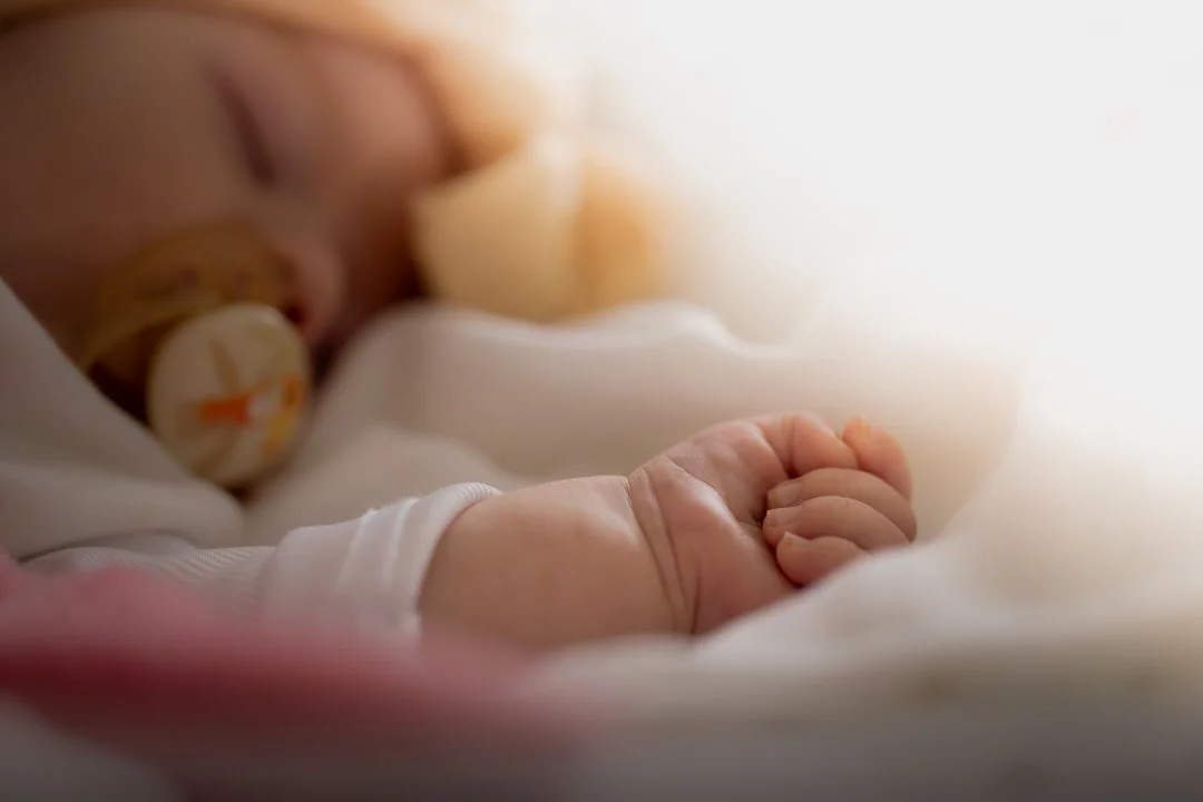 A close-up of a sleeping baby with a pacifier in their mouth, wearing a white long-sleeve outfit, wrapped in soft white blankets.