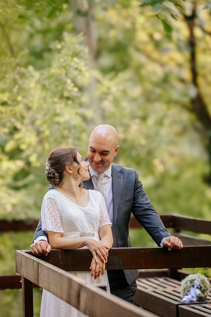 A bride and groom smiling and looking at each other while standing on a wooden bridge outdoors, with trees and green foliage in the background, during their wedding day in Vukovar