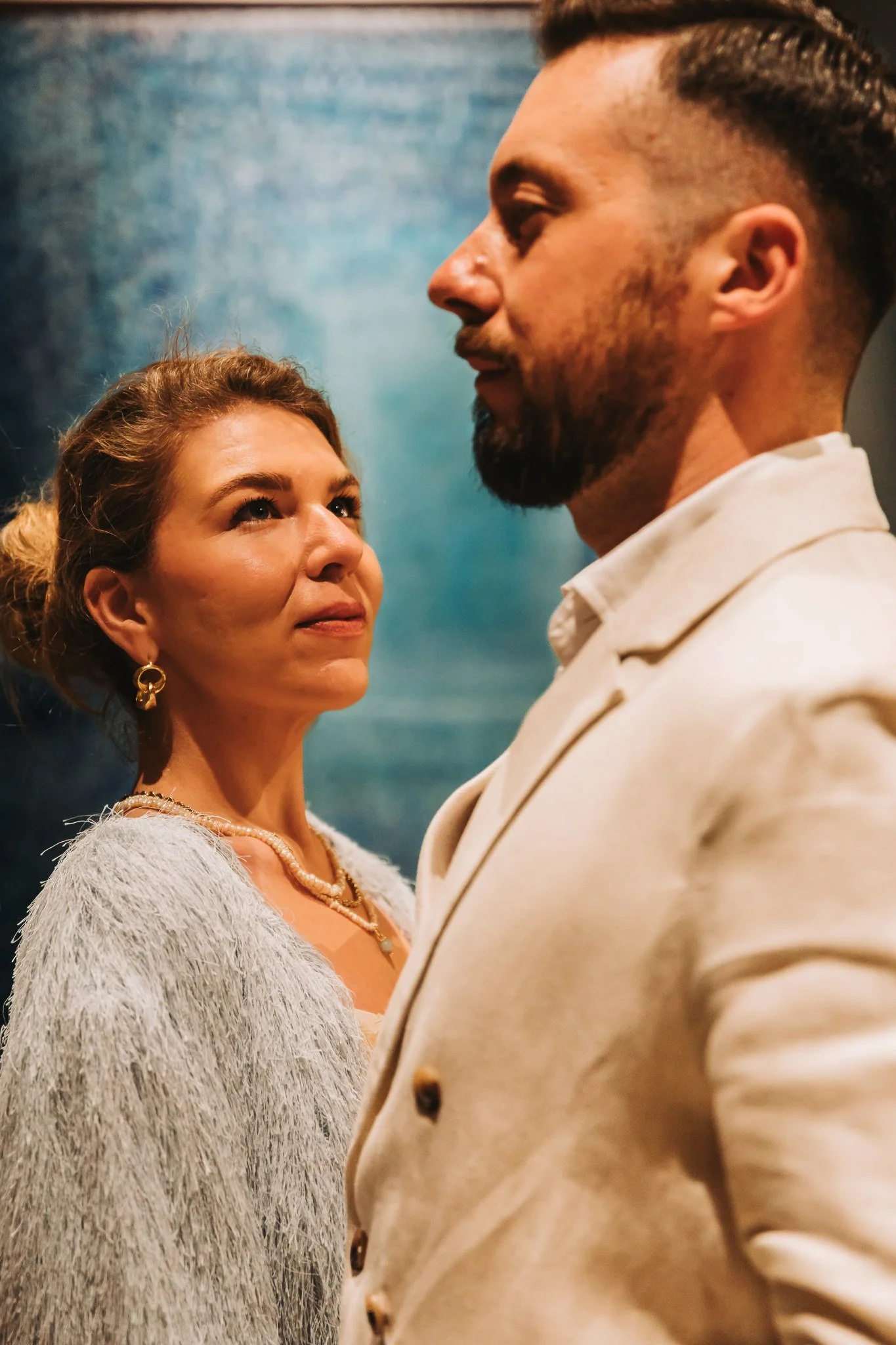 A woman wearing a fluffy gray sweater, pearl necklace, gold earrings, and a pearl bracelet looks up at a man with a beard, wearing a beige blazer, against a blue textured background