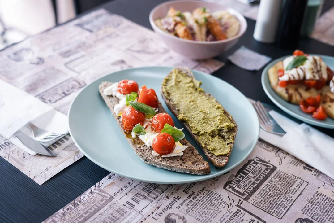 Two slices of toasted bread with toppings: one with cherry tomatoes, basil, and cheese, and the other with a green spread, possibly avocado or guacamole. In the background, there are other plates with food, including pizza and a bowl of salad, on a t