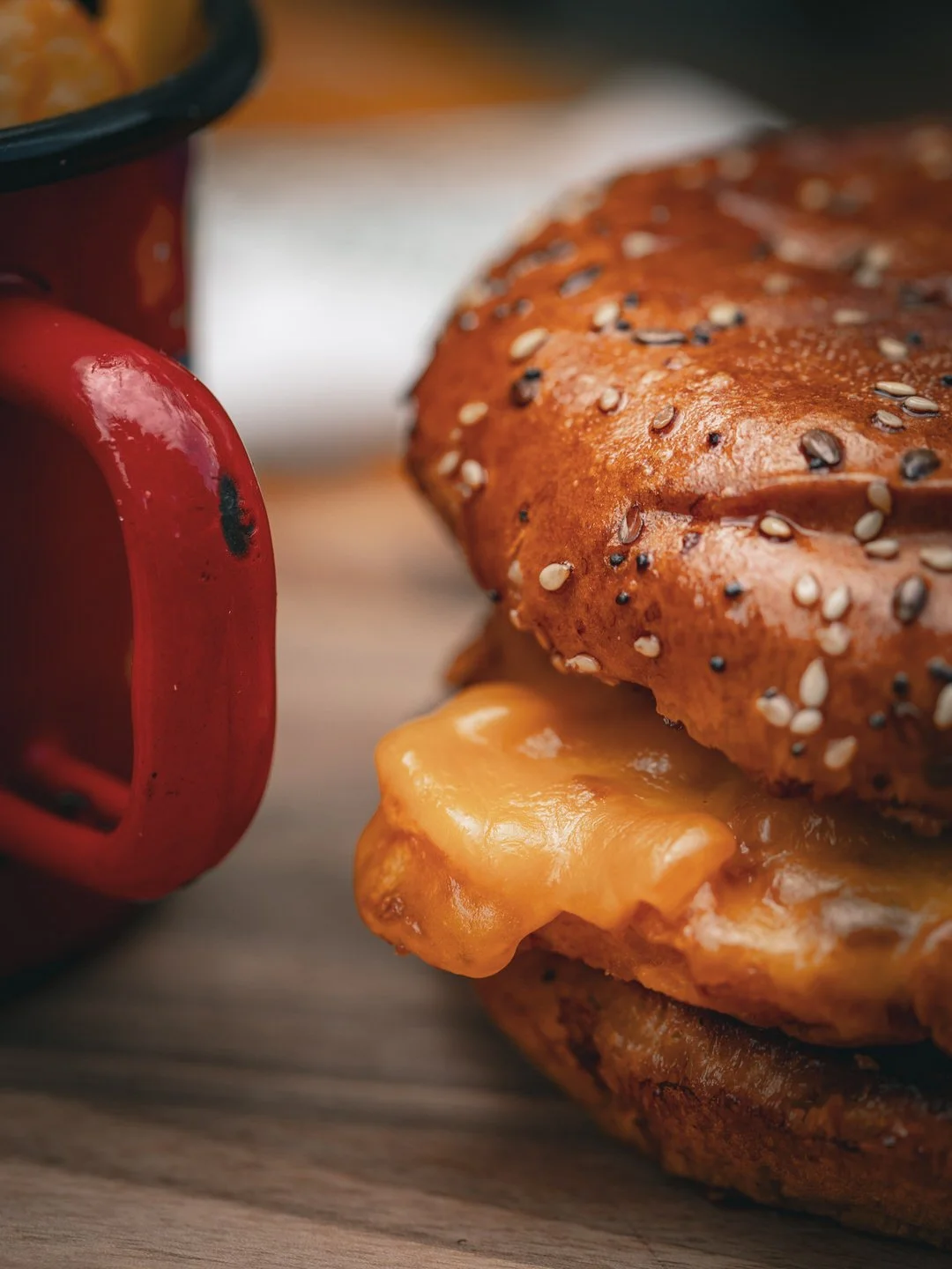 A close-up of a cheeseburger with a sesame seed bun, melted cheese, and a beef patty, next to a red mug on a wooden surface.