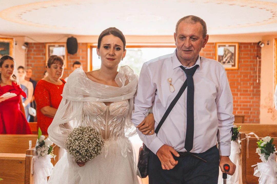 A bride in a wedding dress holding a bouquet of baby's breath walking down the aisle with  her father, during a wedding ceremony. Guests stand on both sides of the aisle in a decorated church with brick walls.