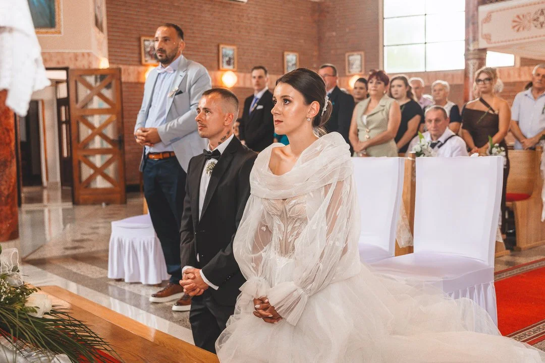 A bride and groom kneeling during their wedding ceremony inside a church, with guests standing behind them.