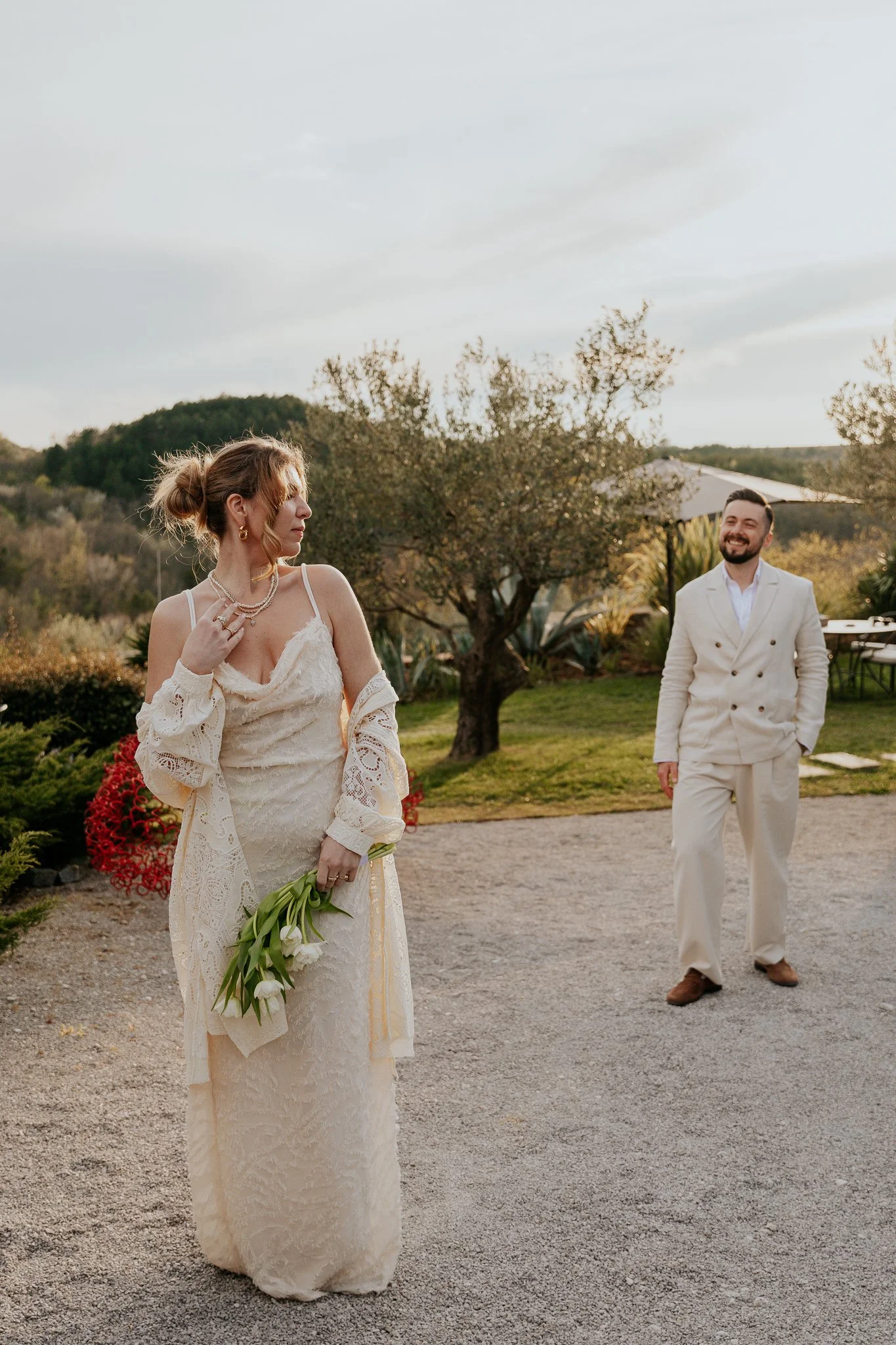 A woman in a white lace dress holding a bouquet of white tulips stands outdoors, looking to her left. A man in a light-colored suit stands to her right, smiling. The background features trees, a building with a white roof, and a hilly landscape under