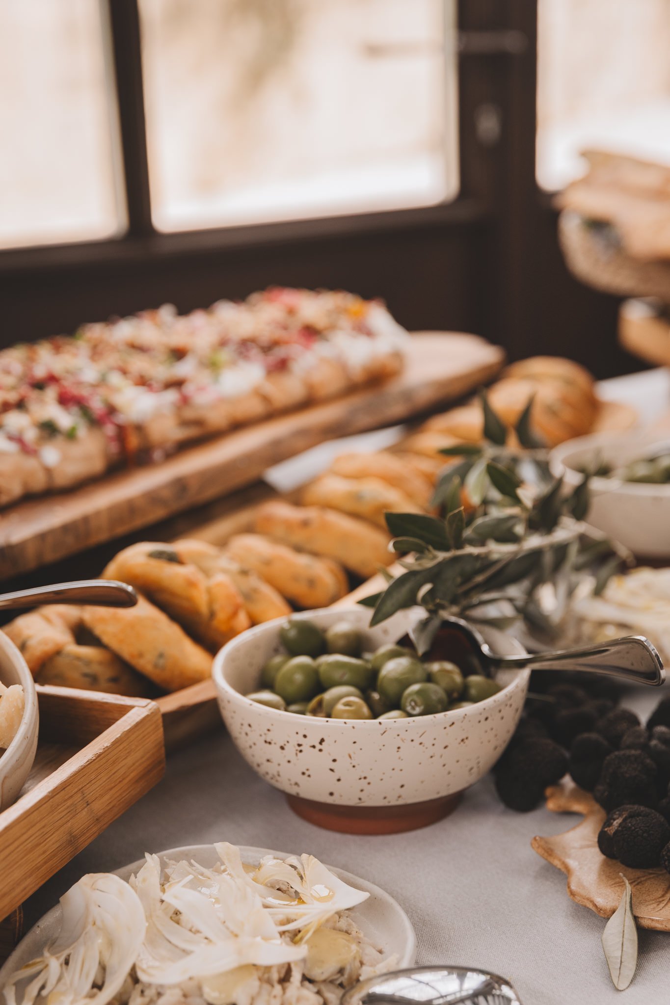 A table with various appetizers and foods, including a bowl of green olives, a plate of sliced cheese, bread rolls, and a tray of what appears to be a flatbread or pizza, with a window in the background.