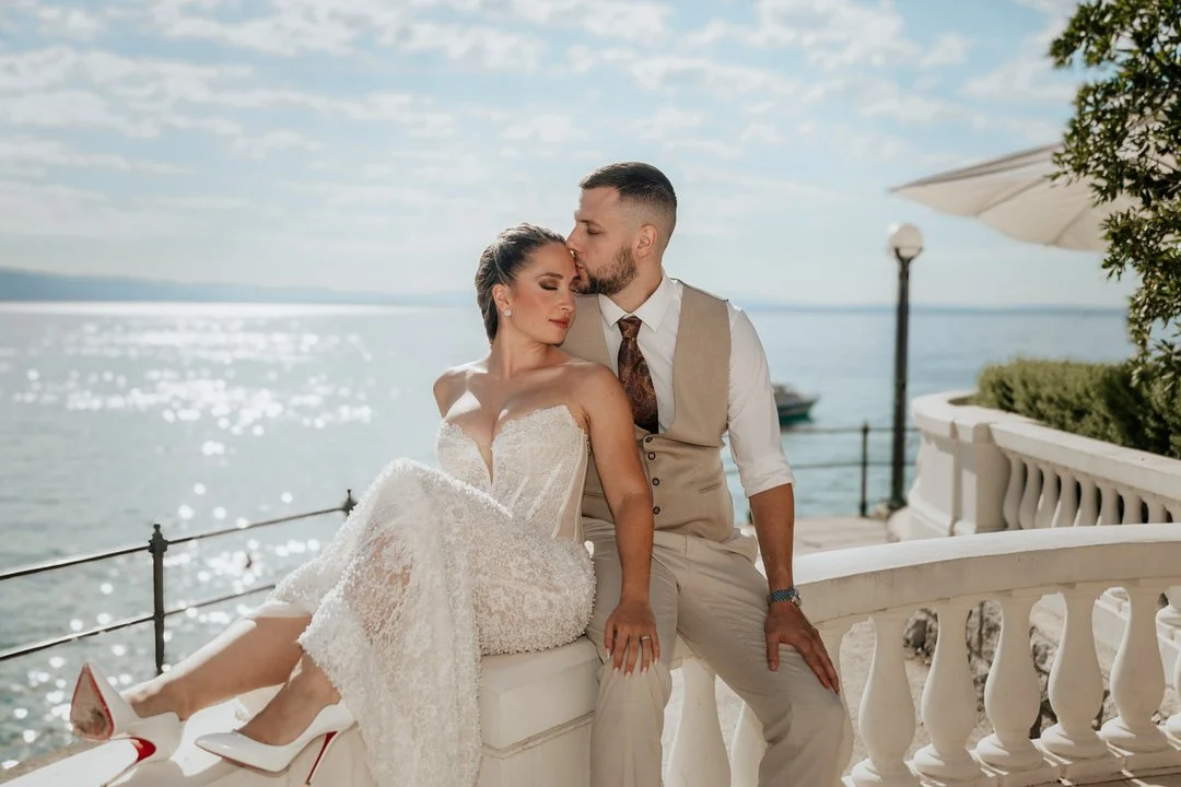 Bride and groom sitting on a white stone balustrade overlooking the sea in front of Hotel Kvarner in Opatija, Kvarner, during their wedding photoshoot.