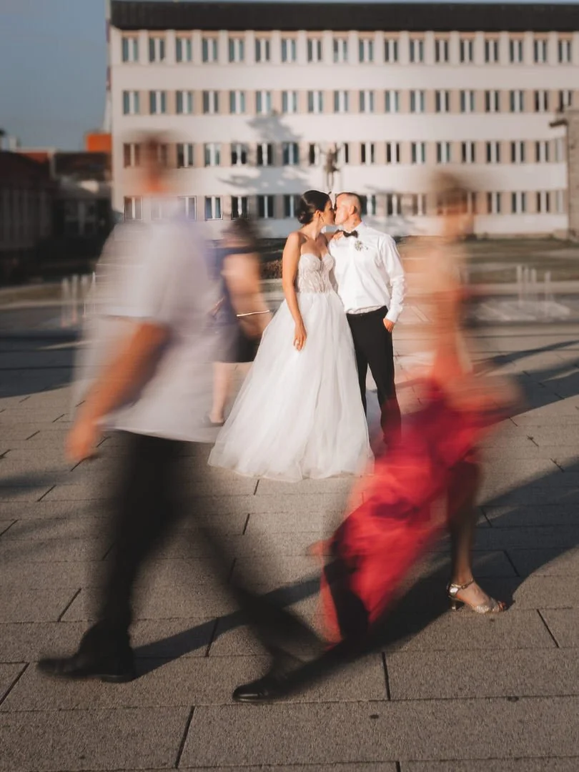 A bride and groom sharing a kiss outdoors on a city street in Našice, with blurred pedestrians walking past in front of them.